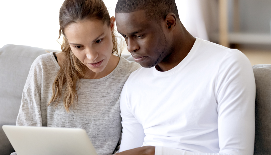 A couple sitting on their couch and looking at a laptop screen together.