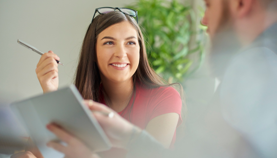 Smiling woman holding a pen and looking at a man while discussing a document, seated indoors with a plant in the background.