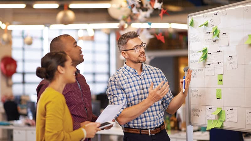 Hispanic male in an office setting working at a whiteboard while collaborating with Black male and Black female coworkers