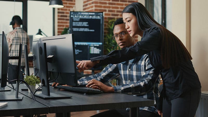 Asian female in an office setting collaborating a black male coworker pointing to a desktop computer monitor