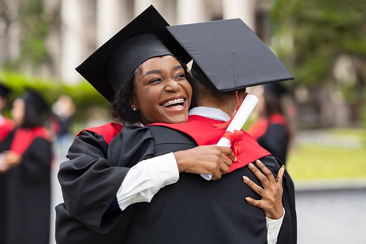 Black female graduate in a cap and gown smiling and hugging another graduate
