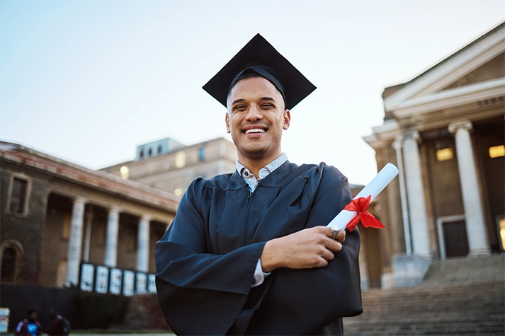 Hispanic male in a cap and gown holding a degree on a college campus 