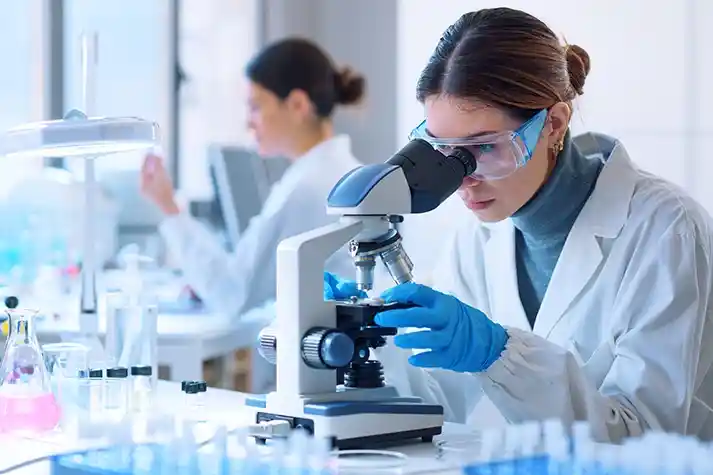 Hispanic female wearing a white lab coat and safety glasses looking into a microscope