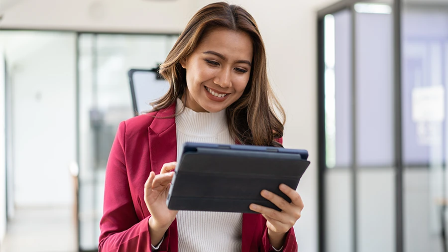 Female Hispanic Chick-fil-A®  Operator standing in a windowed office smiling and looking down at the tablet she is holding.