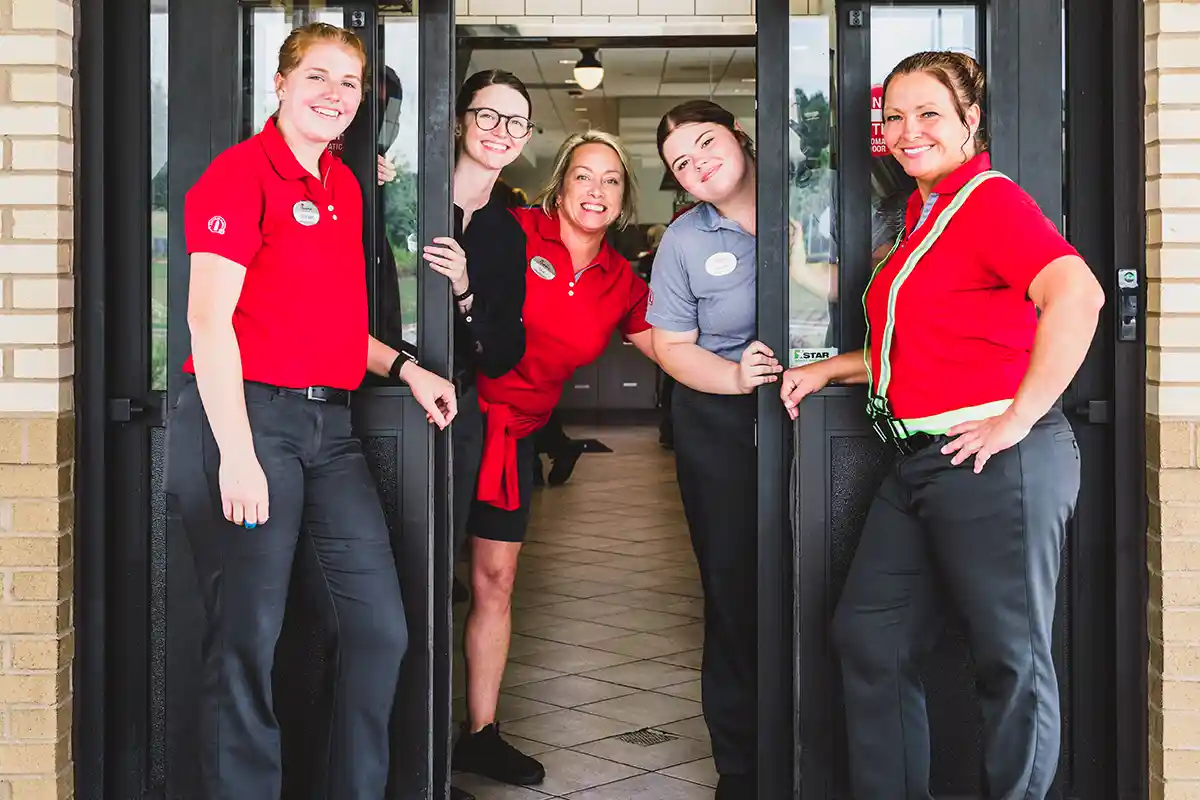 Multi-racial group of female Chick-fil-A® Team Members posing and smiling in the doorway of a Chick-fil-A® restaurant.