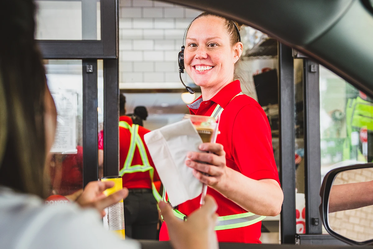 Female Chick-fil-A® Team Member wearing a headset, handing a food order through the drive-through window to a guest in a car.