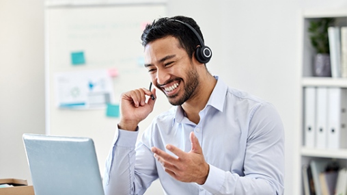 Asian male wearing a headset, taking a call while working on his laptop in an office setting.
