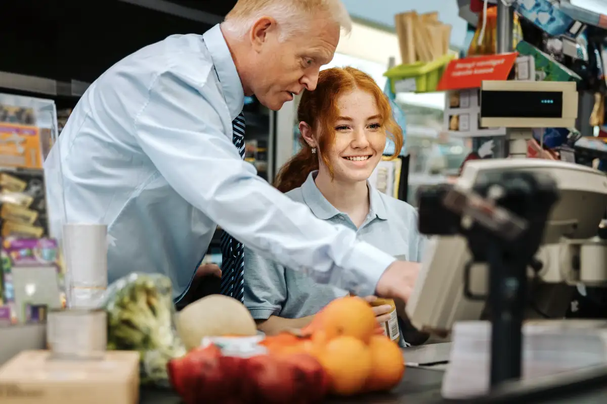 Female grocery store employee with read hair smiling at the checkout stand while a male manager leans over to instruct her on how to scan groceries
