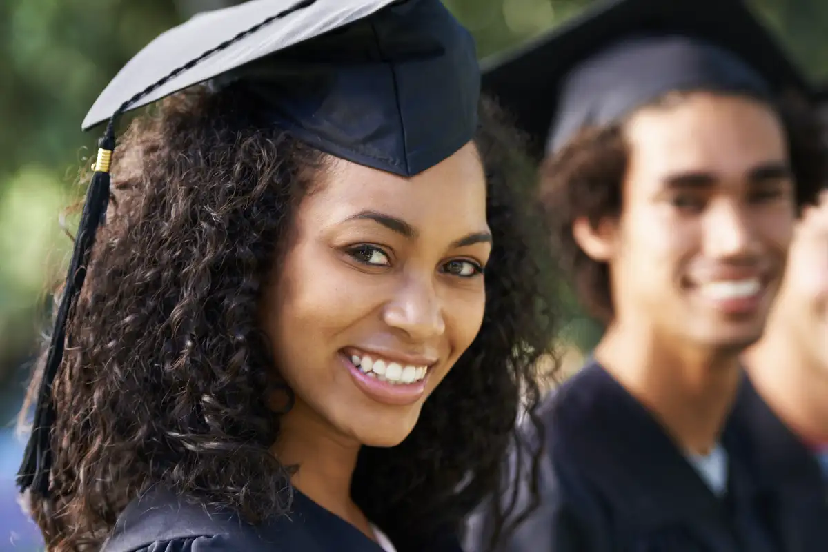 Black female wearing a cap and gown at a college graduation ceremony, sitting next to two other graduates that can be seen in the background