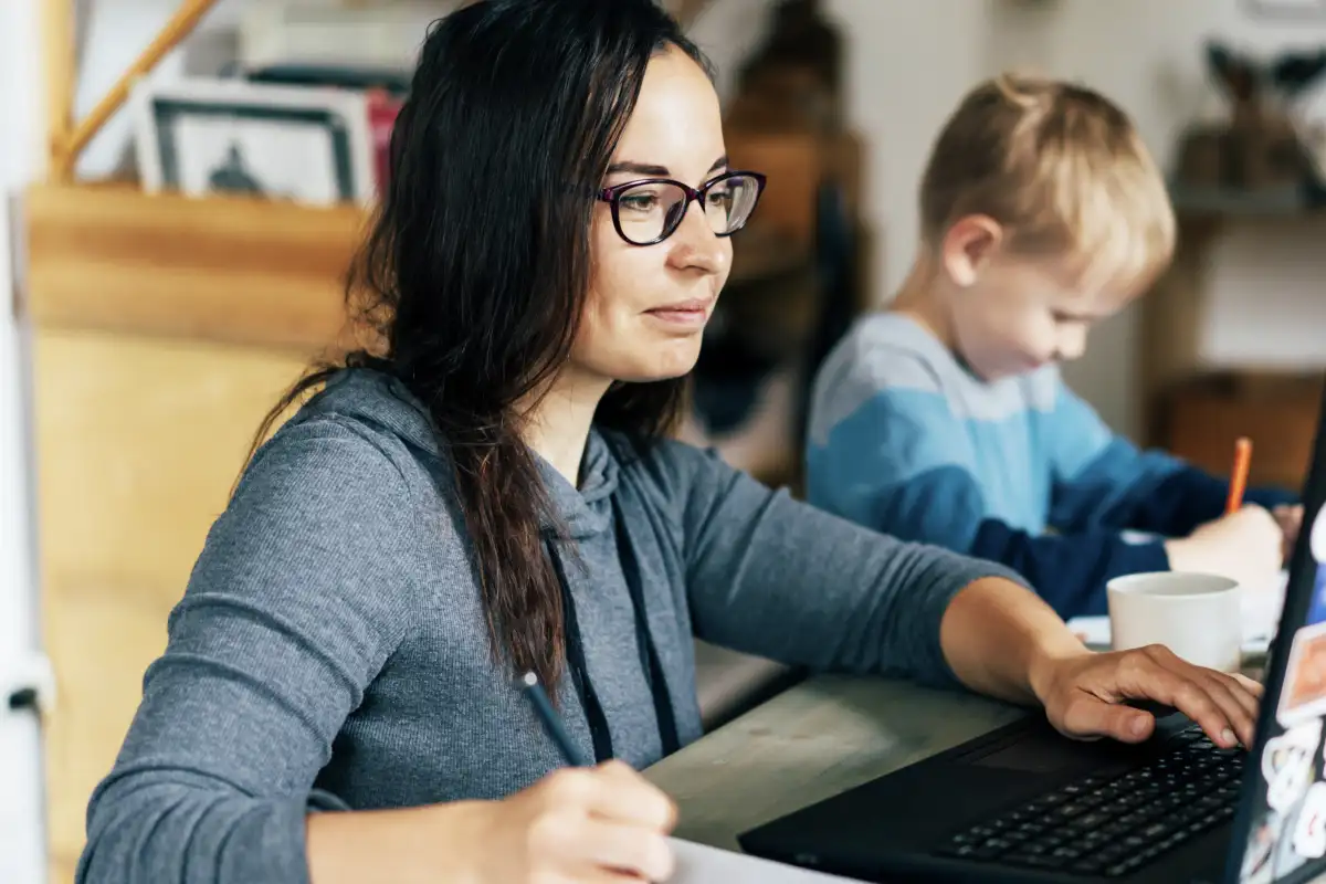 Female working adult learner doing classwork on her laptop at her kitchen table next to her young son who can be seen in the background