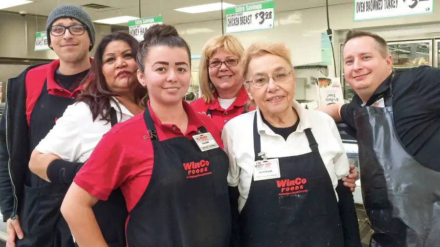 Multi-racial and multi-generational group of Winco employees posing for a picture in front of a deli case inside a Winco