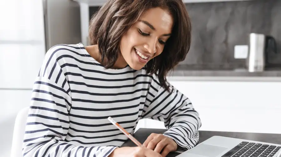 Black female student sits in front of an open laptop in her kitchen while writing notes on paper with a pencil