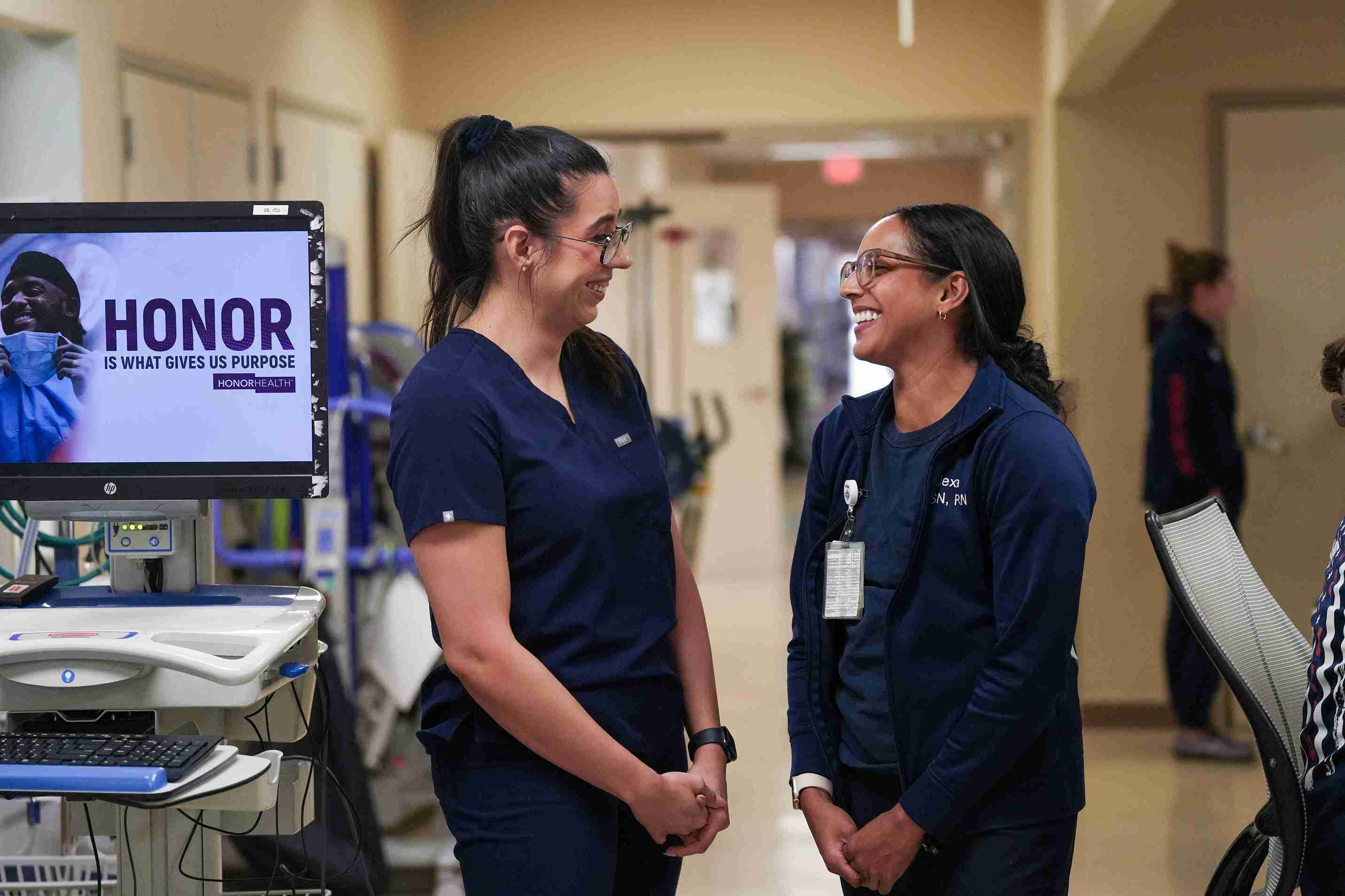 Two female Honor Health employees wearing scrubs standing in a hospital hallway smiling at each other