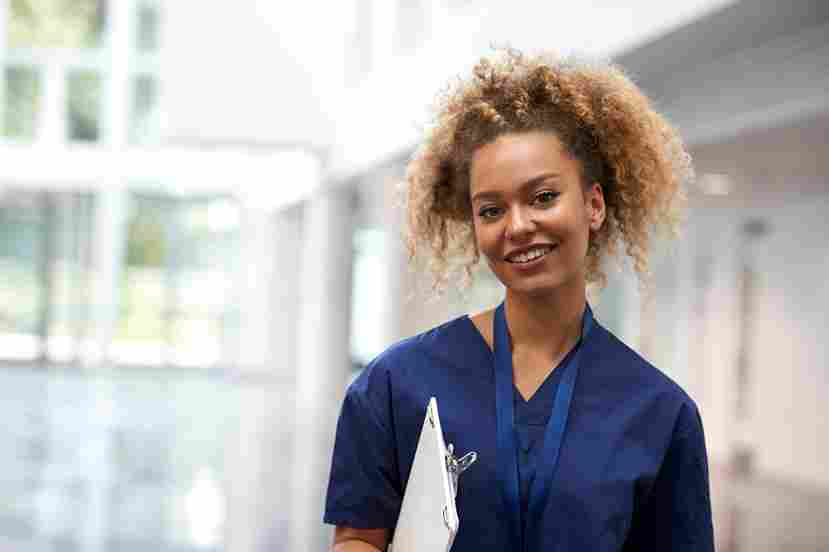 A young woman in blue scrubs smiles confidently while standing in a bright, modern healthcare facility.
