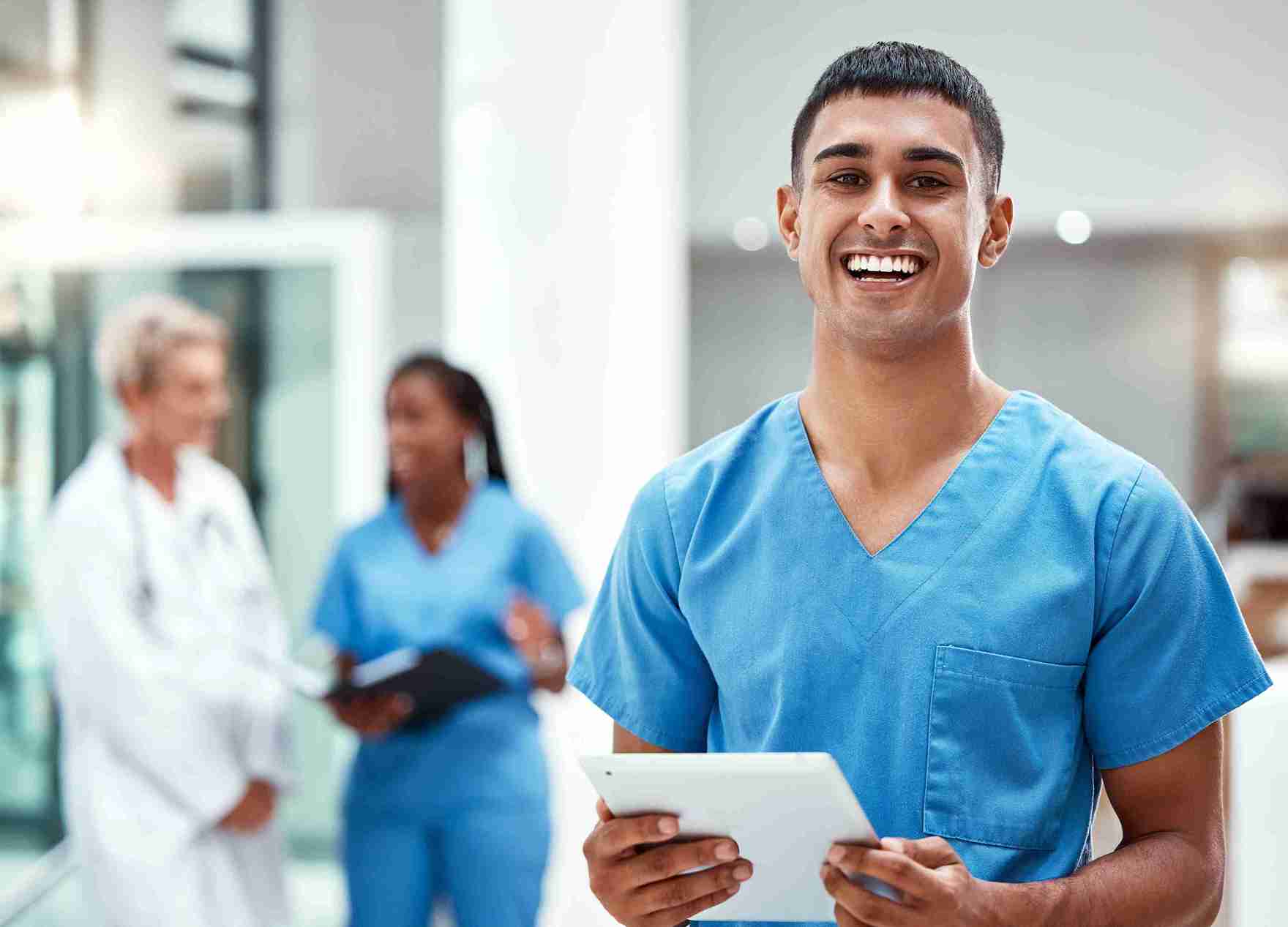 A smiling health care worker in blue scrubs stands in a bright medical facility, with two colleagues conversing in the background.