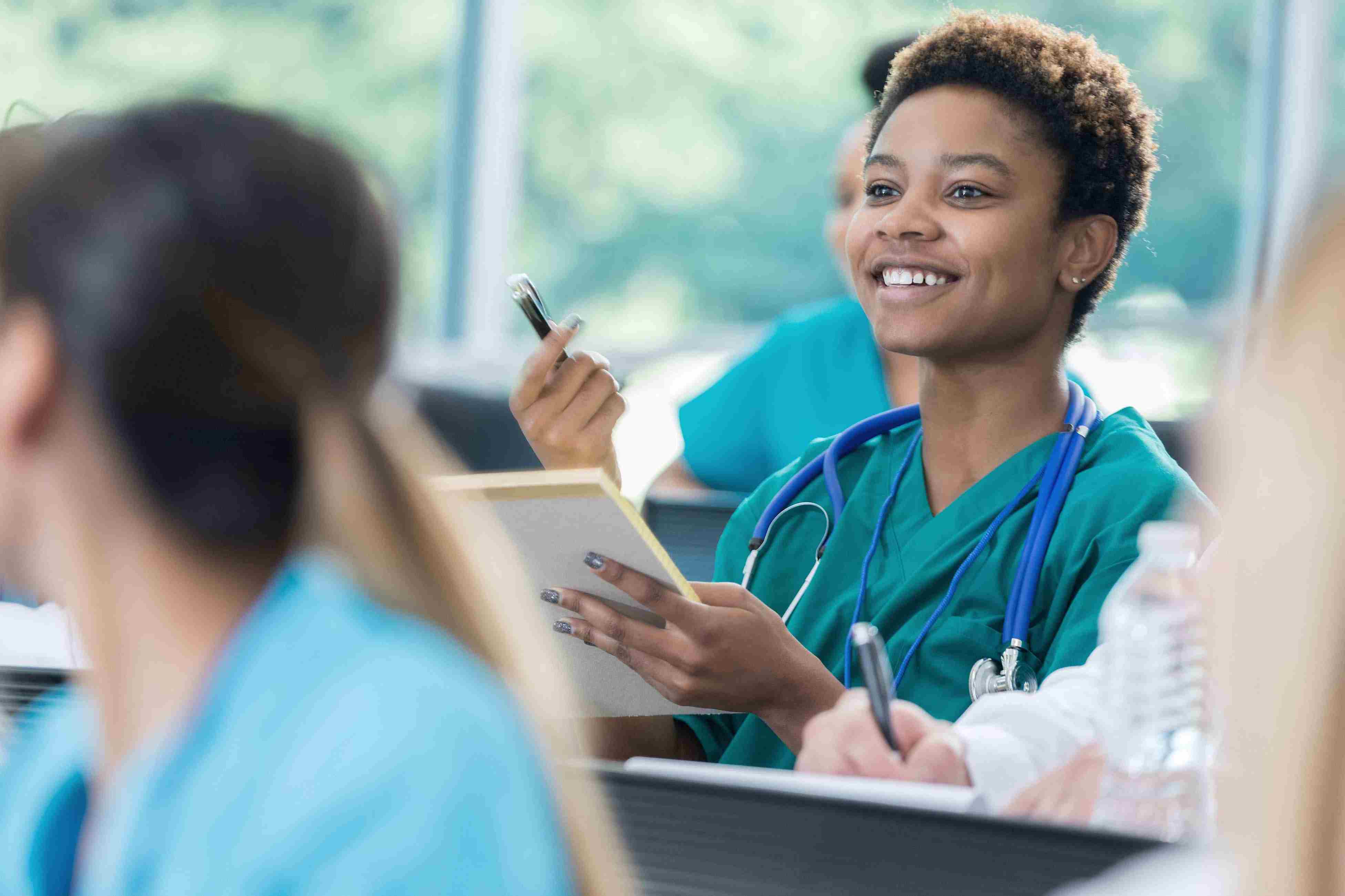 A smiling health care professional in scrubs holds a pen and notebook during a classroom or training session, with others seated nearby.
