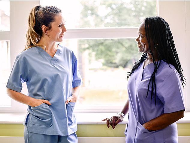 Two female nurses in scrubs standing in front of a window smiling at one another